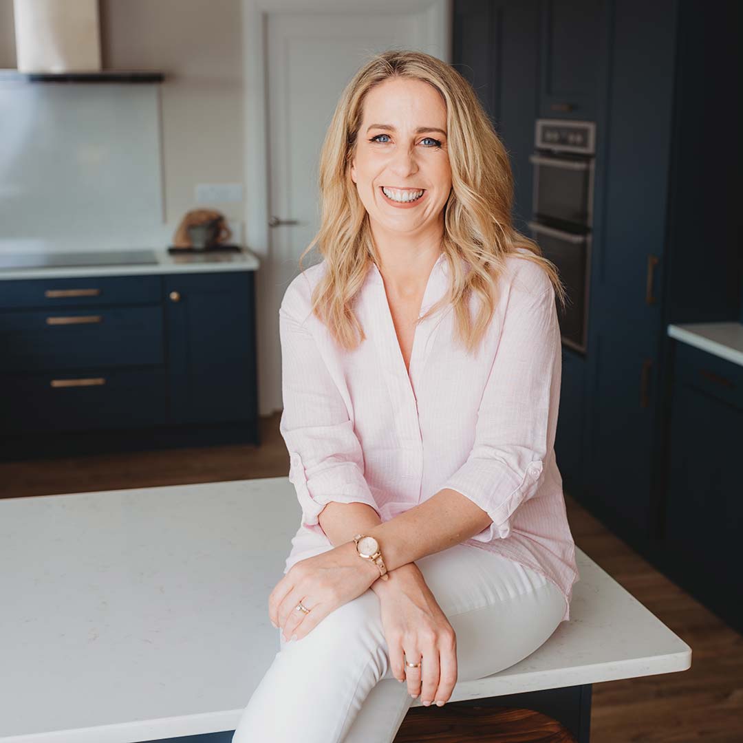 blonde woman sitting on kitchen counter top wearing pink shirt and white jeans