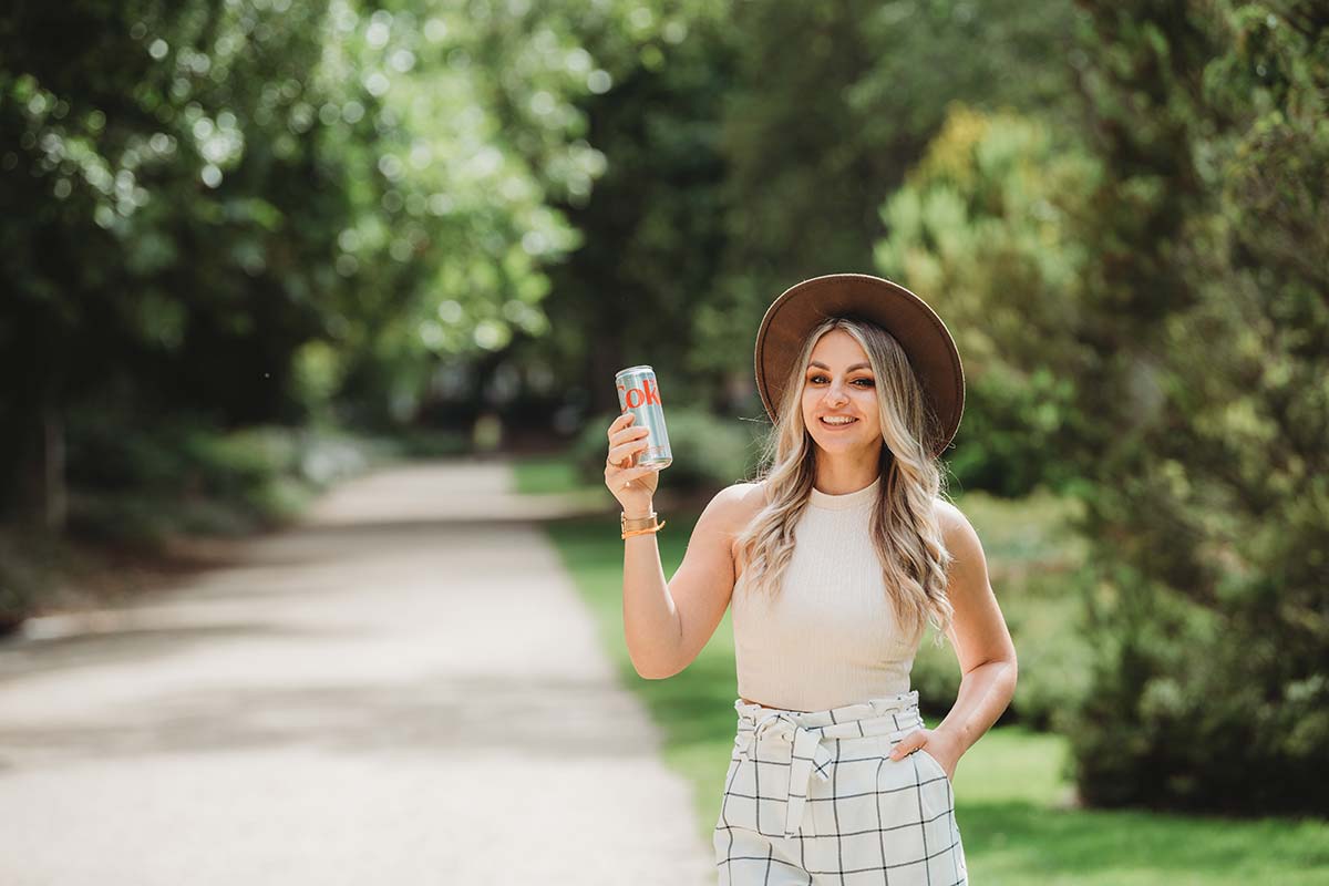 female holding can of Diet Coke with hand in pocket smiling at camera in the park