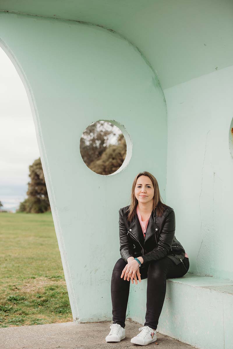 woman wearing black jeans, black leather jacket and sitting in beach front green shelter