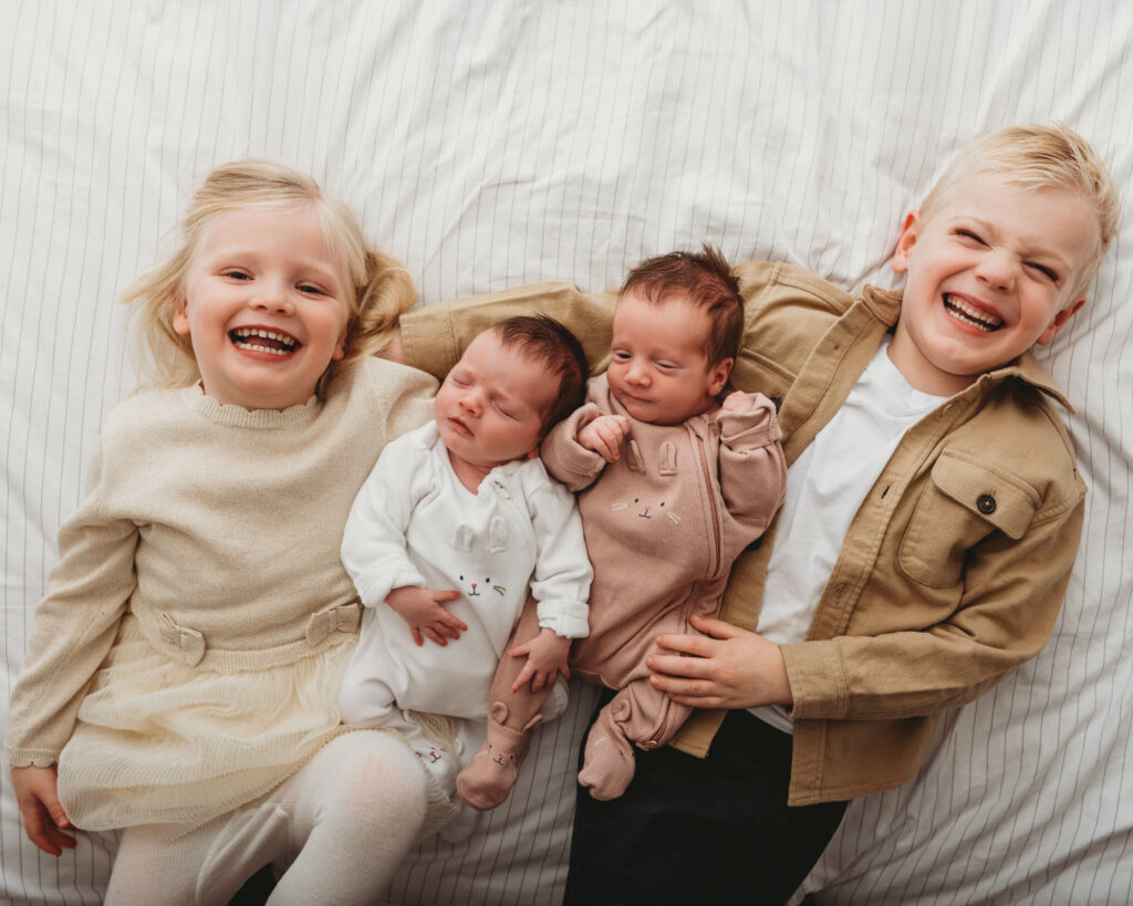Four siblings lying on bed, older brother and sister are laughing while holding their new twin sisters.