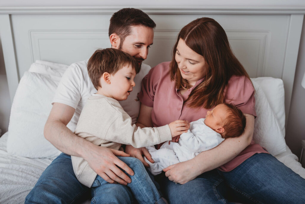 Family of four cuddled together on parents bed, big brother holding his new baby sisters hand.