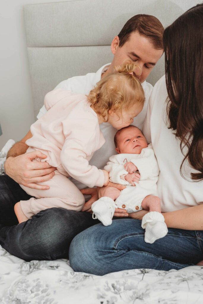Parents holding their toddler and baby while the toddler gives the baby a kiss on the forehead.